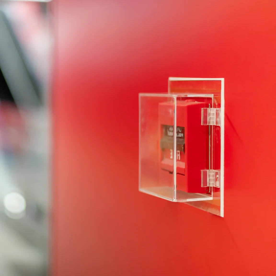Eine Nahaufnahme eines roten Feueralarms in einem transparenten Gehäuse an einer Wand. Der Hintergrund ist unscharf, mit einer modernen, roten Wand, die das Sicherheitsgerät hervorhebt.