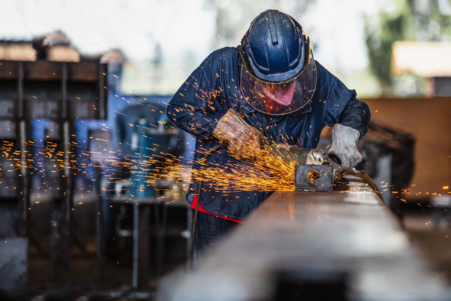 Ein Handwerker schweißt an einem Metallstück. Funken fliegen aus dem Schweißbereich, während er mit Schutzausrüstung wie einem Helm und Handschuhen arbeitet. Der Fokus liegt auf der Funkenbildung, die das Bild dynamisch wirken lässt.