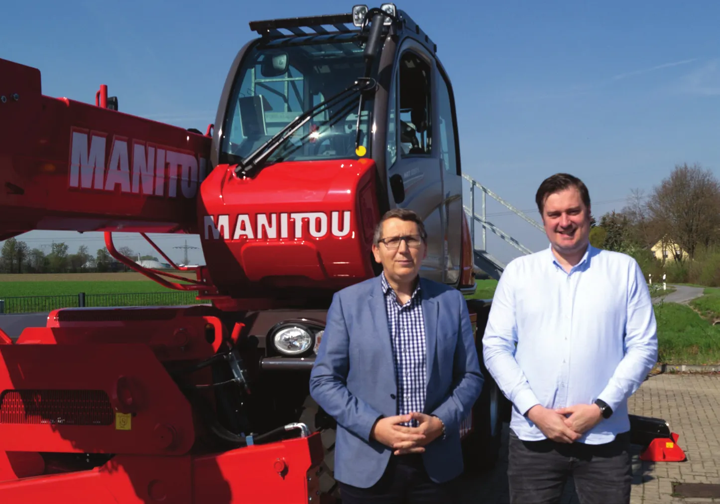 Das Bild zeigt zwei Männer, die vor einer großen roten Maschine stehen, vermutlich ein Bau- oder landwirtschaftliches Gerät, auf dem deutlich der Name „MANITOU“ zu sehen ist. Die Szene spielt sich im Freien an einem sonnigen Tag ab, im Hintergrund sind grüne Felder zu erkennen. Die Männer tragen legere Businesskleidung.