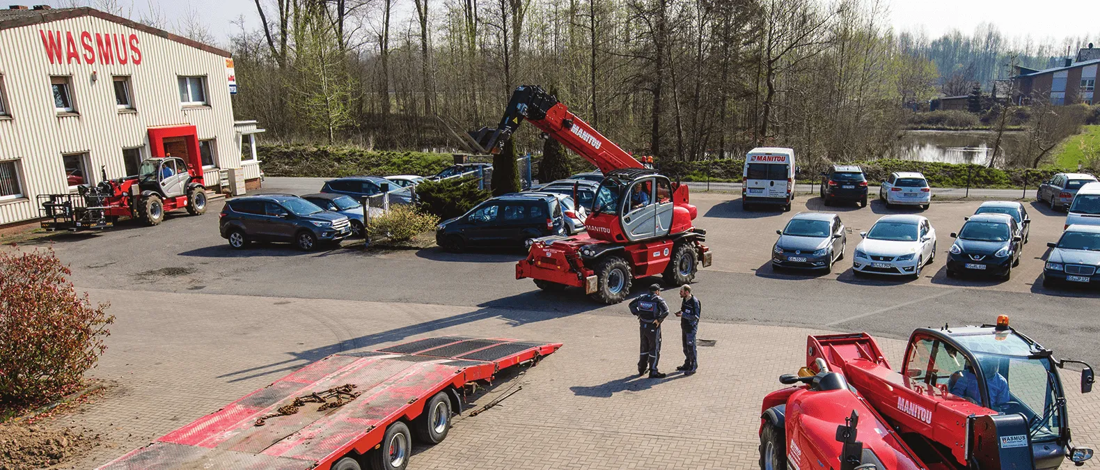 Das Bild zeigt einen Parkplatz vor einem Gebäude, auf dem der Name „WASMUS“ gut sichtbar angebracht ist. Es sind mehrere Fahrzeuge geparkt, darunter Autos und eine große rote Maschine – vermutlich ein Bau- oder landwirtschaftliches Gerät mit einem kranartigen Ausleger. Zwei Personen in Arbeitskleidung stehen in der Nähe der Maschine, möglicherweise in ein Gespräch vertieft oder bei einer Begutachtung. Im Hintergrund sind Bäume und ein Gewässer zu sehen, was auf eine naturnahe Umgebung hinweist.