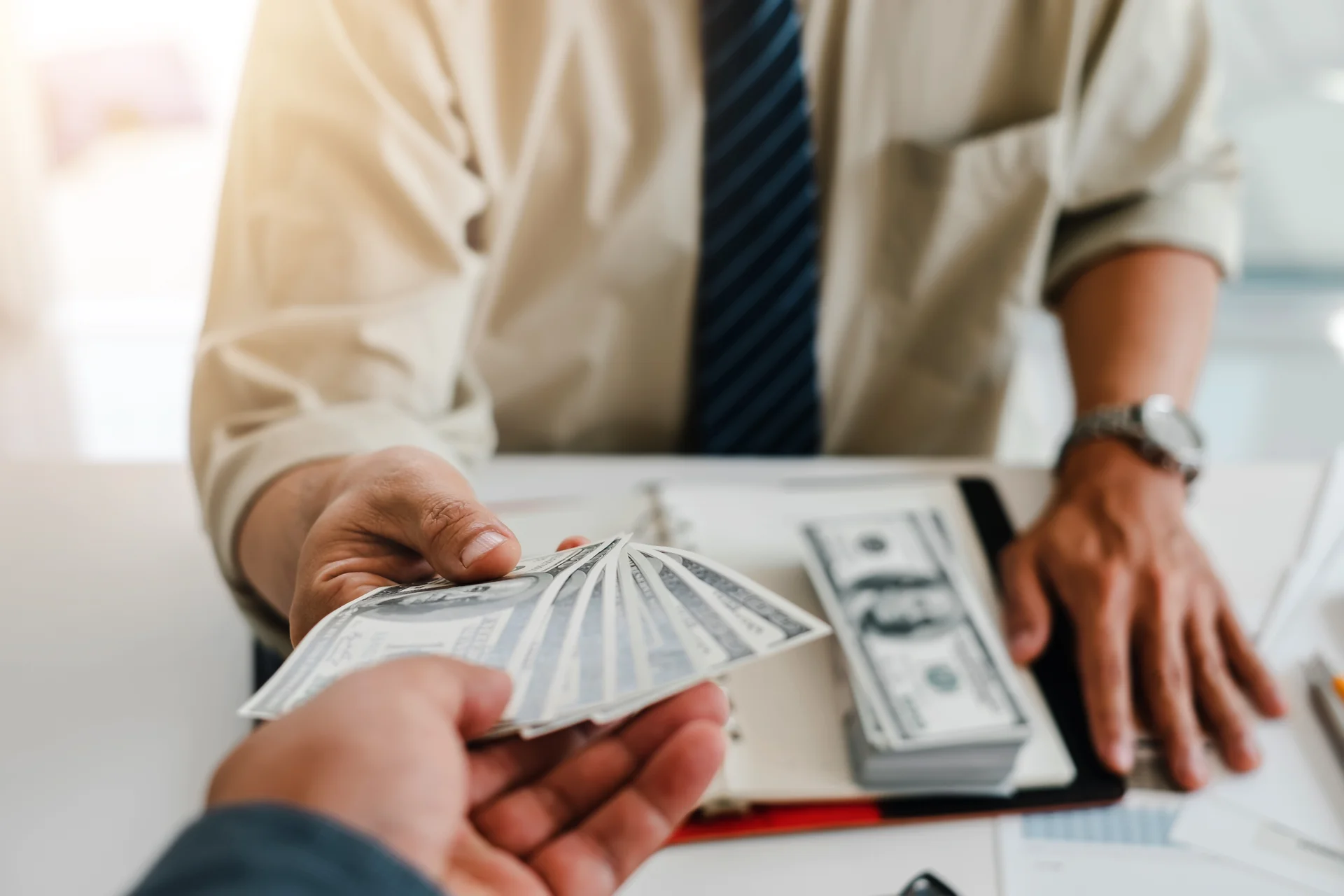 Ein Geschäftsmann in Hemd und Krawatte übergibt einem anderen Menschen mehrere 100-Dollar-Scheine, im Hintergrund liegen weitere Geldscheine auf einem Schreibtisch.